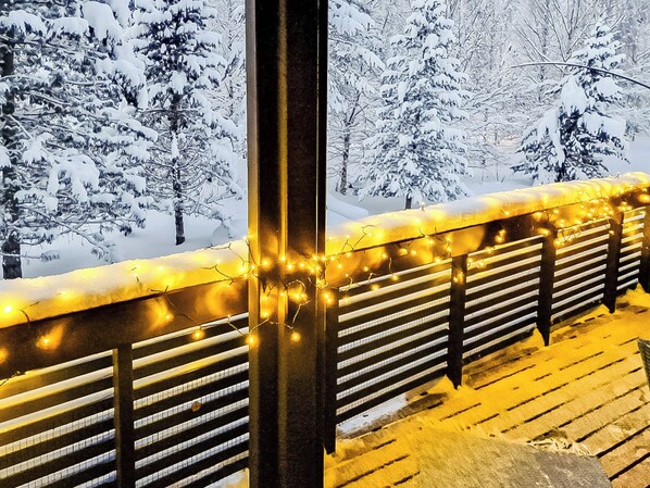A snow-covered landscape with a wooden deck and railing, illuminated by string lights.
