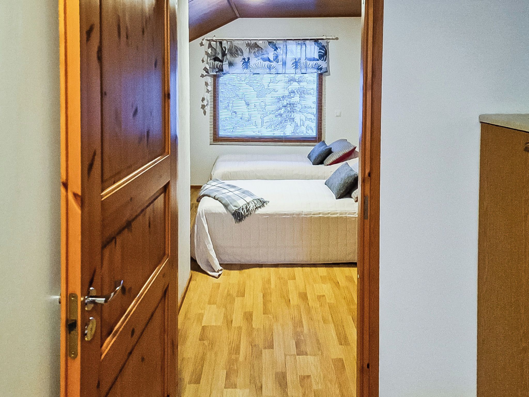 Una habitación estrecha con una puerta de madera, una cama con almohadas, una ventana con un mapa mundial y un suelo de madera.