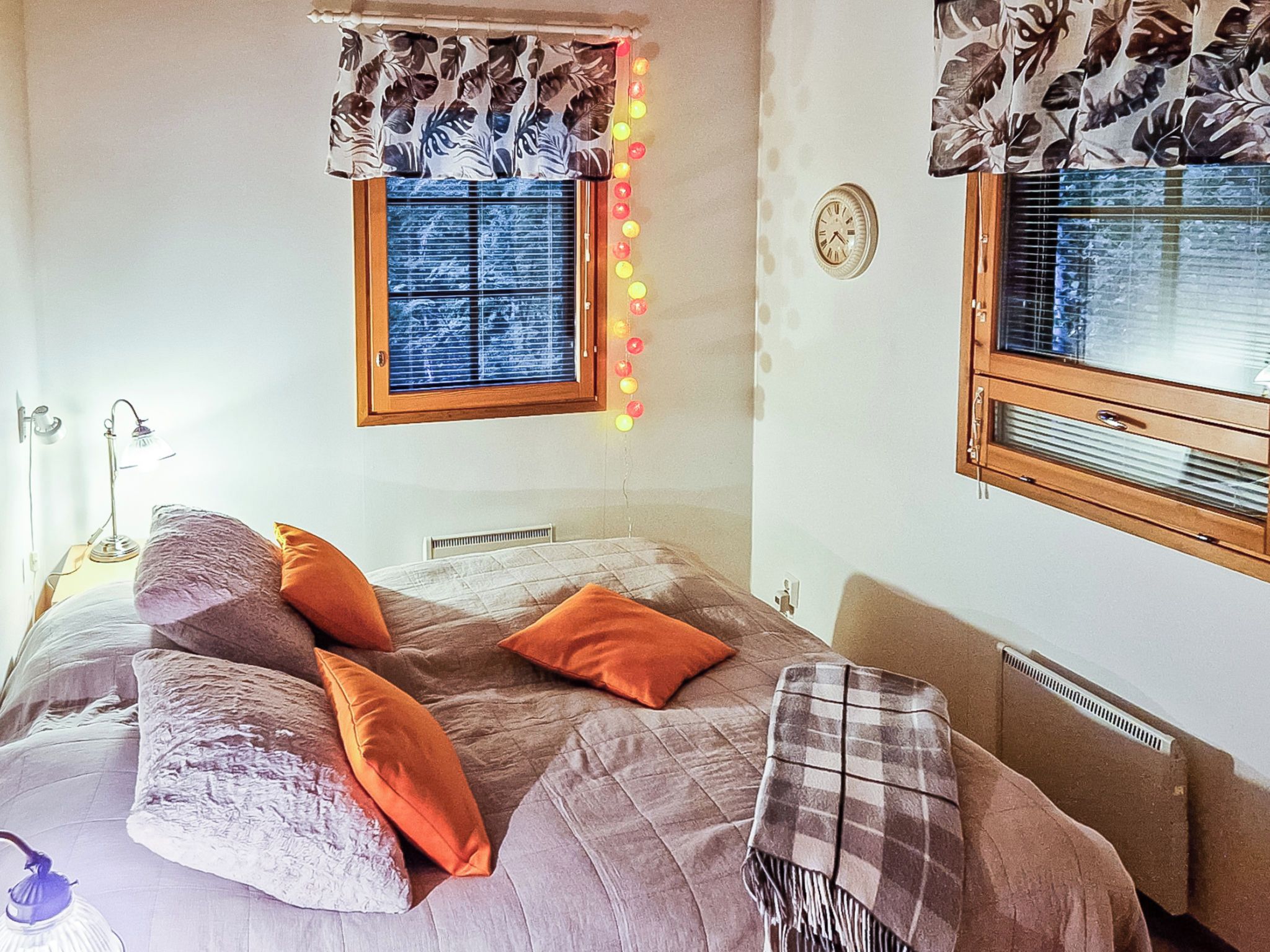 A bedroom with a bed, pillows, a window with blinds, a wall clock, and a radiator.