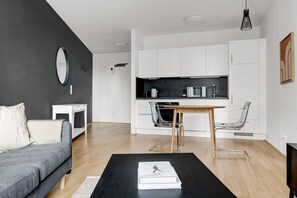 Uma sala moderna com um sofá cinza, uma mesa de centro preta com um livro, uma mesa de jantar de madeira com cadeiras e uma área de cozinha com armários e eletrodomésticos brancos.