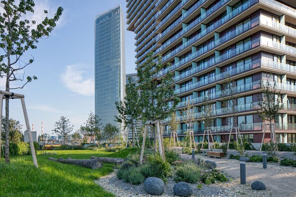 A modern building with multiple floors and balconies, surrounded by landscaped gardens with trees and a gravel path.