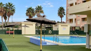 Ein Swimmingpool mit blauer Fliesenleiste, umgeben von künstlichem Rasen, Palmen und einem Gebäude mit Balkonen.