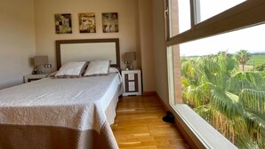 Ein Schlafzimmer mit einem großen Bett, zwei Nachttischen und Blick auf Palmen durch ein Fenster.