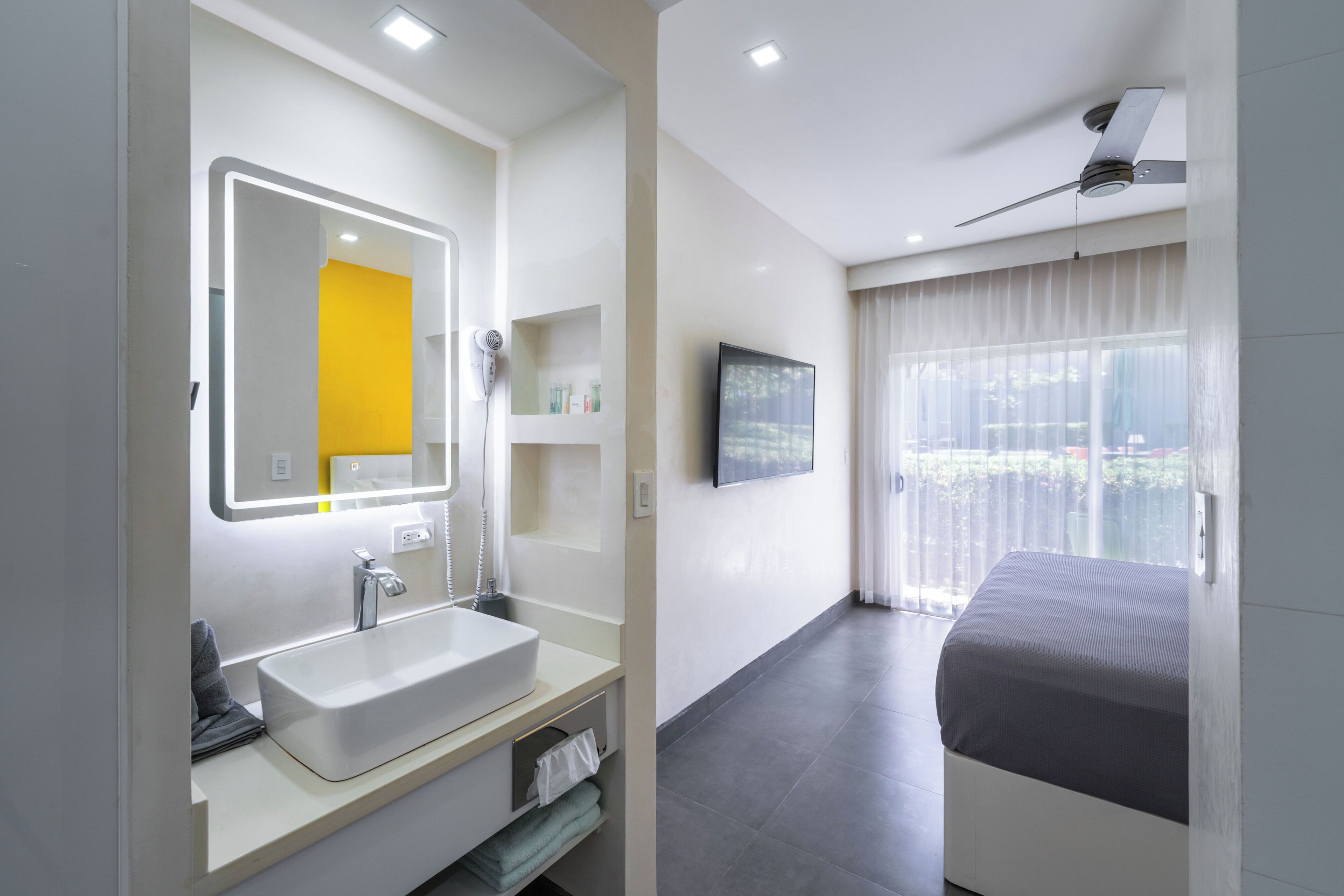 Une salle de bain moderne avec un grand miroir, un lavabo et une douche. Il y a un lit avec vue sur l’extérieur.