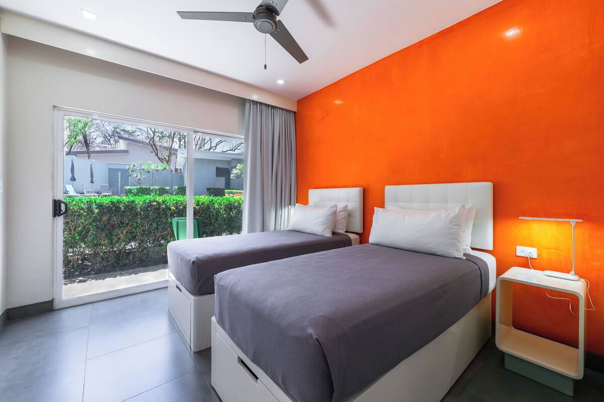 A modern hotel room with two beds, a ceiling fan, and a view of the outdoors.