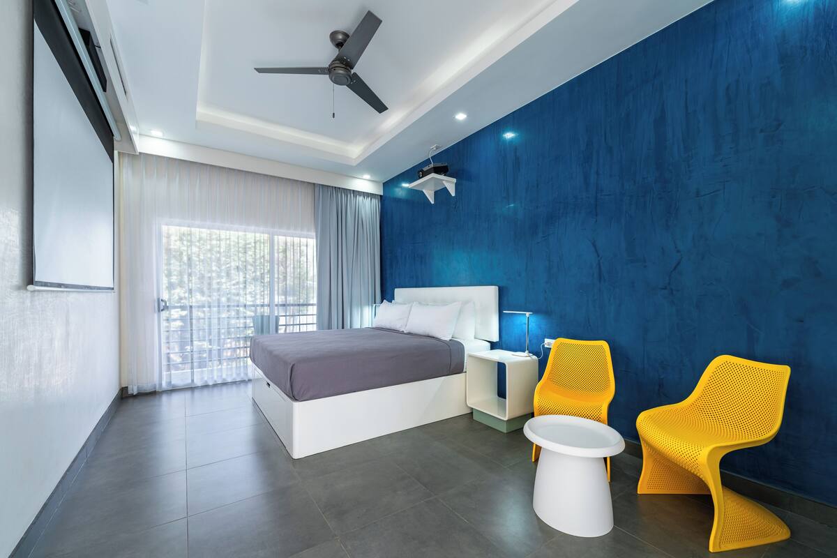 A modern hotel room with a bed, bedside tables, a ceiling fan, a window with curtains, and two yellow chairs.