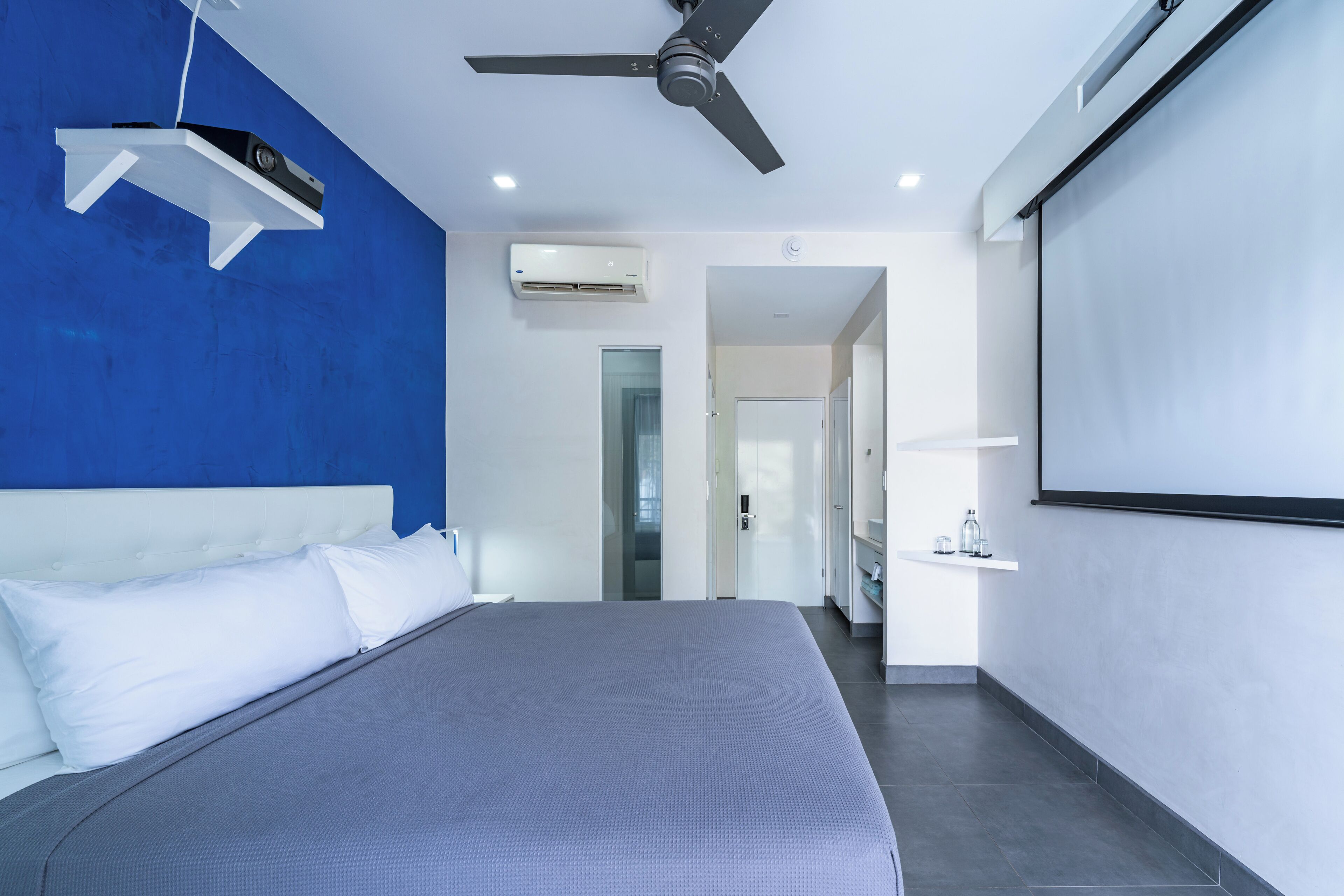 Une chambre d’hôtel moderne dotée d’un grand lit, d’un ventilateur de plafond, d’un appareil de climatisation, d’un écran de projection et d’une salle de bain avec douche et lavabo.