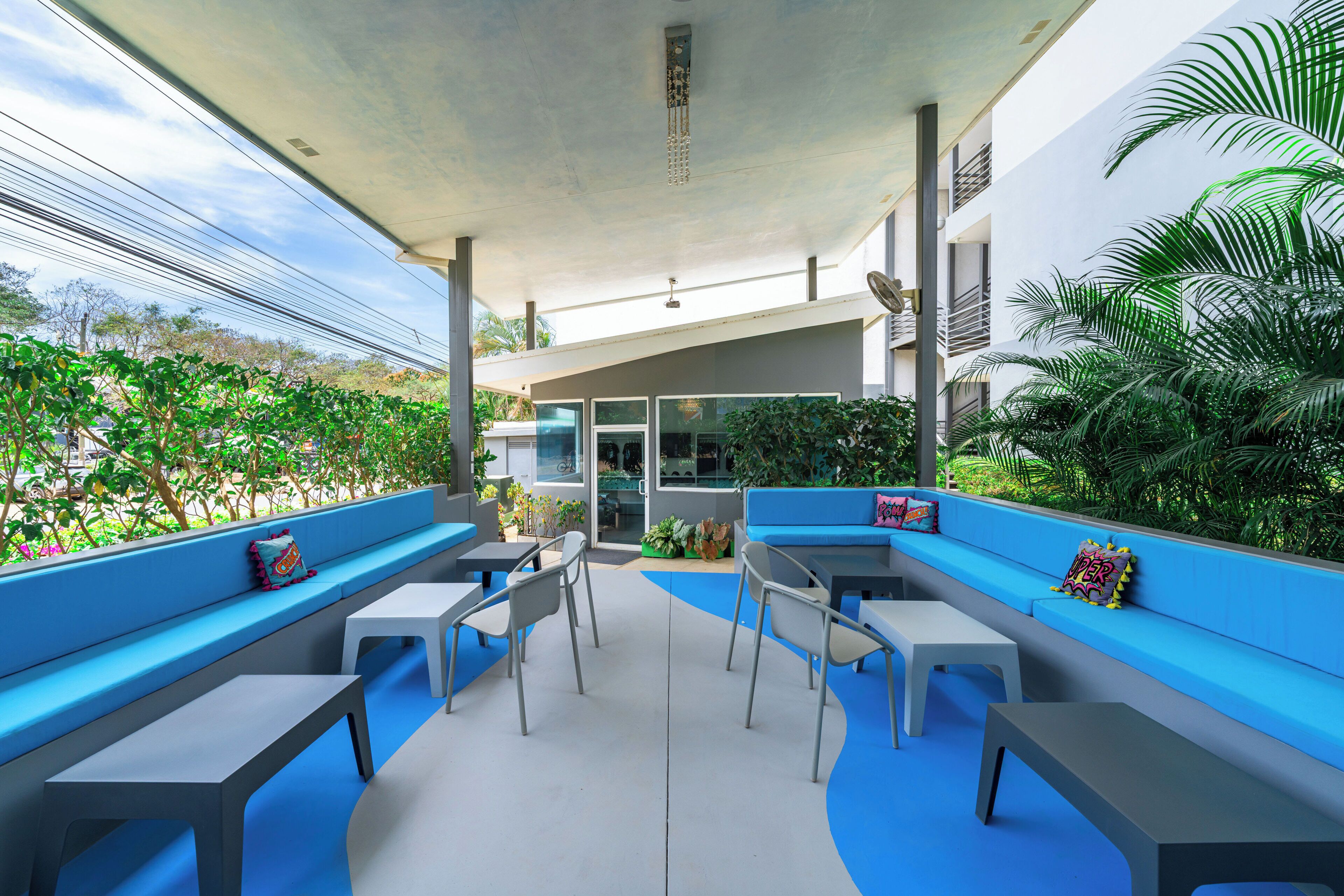Un área de estar al aire libre con bancos y mesas azules, rodeada de vegetación y un edificio moderno al fondo.