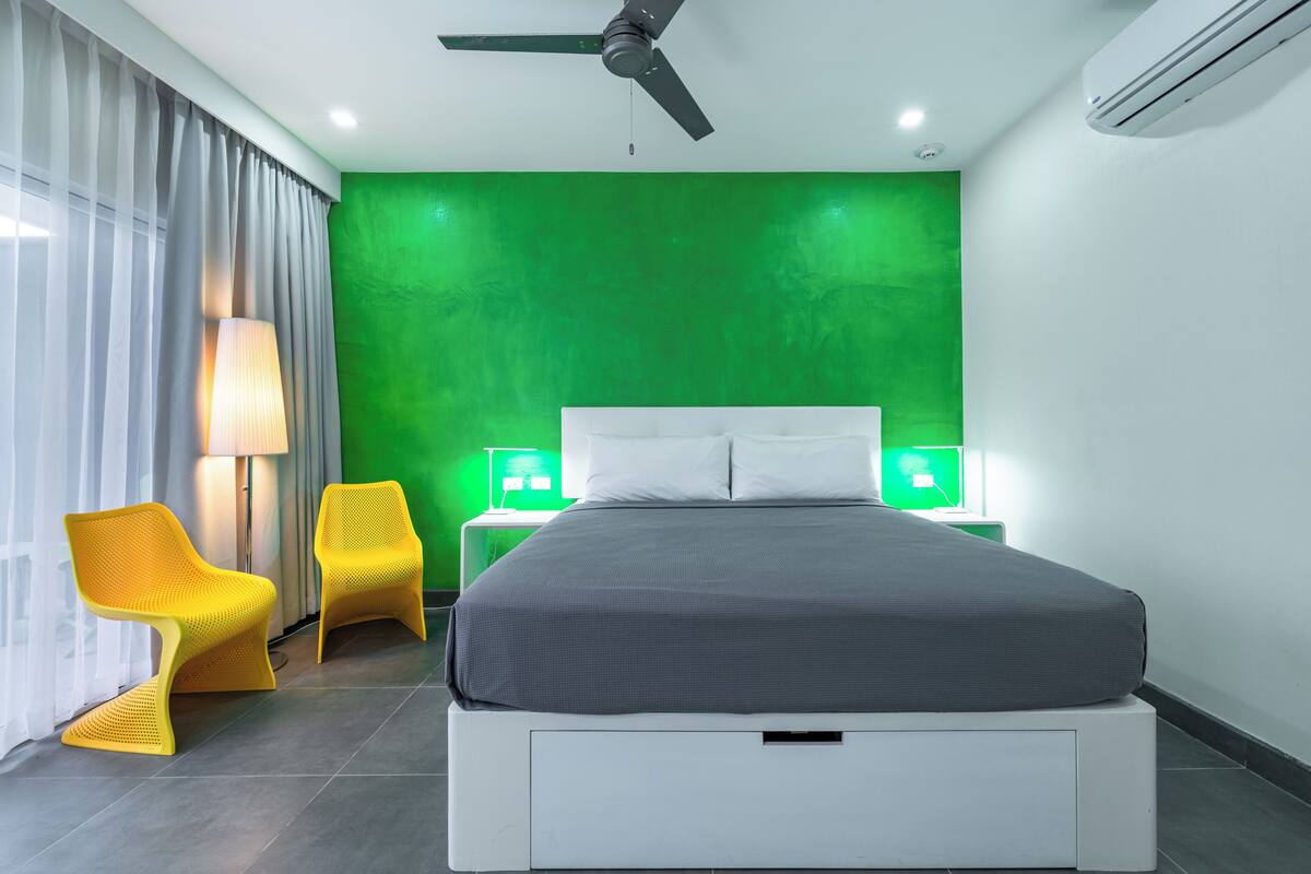 A modern hotel room with a bed, two chairs, a lamp, and a green accent wall.