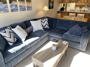 A modern living room with a grey sectional sofa, a wooden coffee table, and a kitchen area in the background.