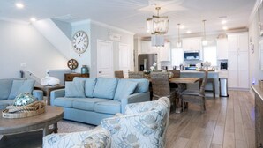Amplia sala de estar con un sofá azul claro, un sillón estampado y una mesa de centro de madera. Se observa un comedor con sillas de mimbre y una mesa de madera. El área de la cocina está equipada con electrodomésticos modernos y armarios blancos.