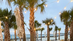 Un patio frente a la playa con mesas de picnic blancas y luces colgantes, con vistas al océano.
