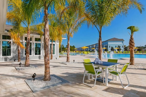 A poolside area with a table and chairs, palm trees, and a building in the background.