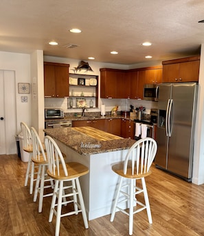 A modern kitchen with wooden cabinets, a stainless steel refrigerator, a built-in oven, and a granite countertop. There are four white bar stools around the island.