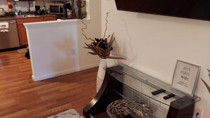 Una sala de estar moderna con un televisor de pantalla plana montado en la pared, una mesa de centro de vidrio con objetos decorativos, y una zona de cocina al fondo.
