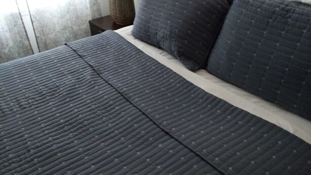Una cama bien tendida con colcha y almohadas azul oscuro, una lámpara de noche y una ventana con cortinas traslúcidas.