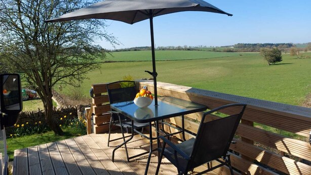 A deck with a table and chairs, an umbrella, and a view of a green field. - Large Static Motorhome-Stunning Countryside Views (Cirencester)