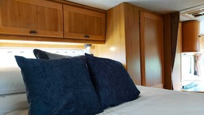 A bed with two dark pillows and wooden cabinets above. - Large Static Motorhome-Stunning Countryside Views (Cirencester)