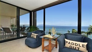 Un salon moderne doté d’une grande baie vitrée donnant sur l’océan, de deux fauteuils bleus ornés de coussins à motifs de vagues, d’une table basse en bois et d’un coin repas avec une table et des chaises.