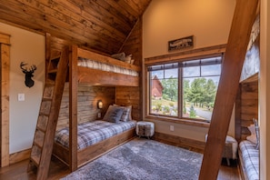 A rustic cabin room with a loft bed, a ladder, a large window with a view of a red barn, and a mounted deer head on the wall.