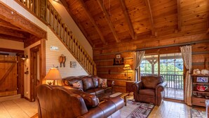 Interior de una cabaña de troncos con escalera de madera, mobiliario de cuero y un ventanal con vistas al exterior.