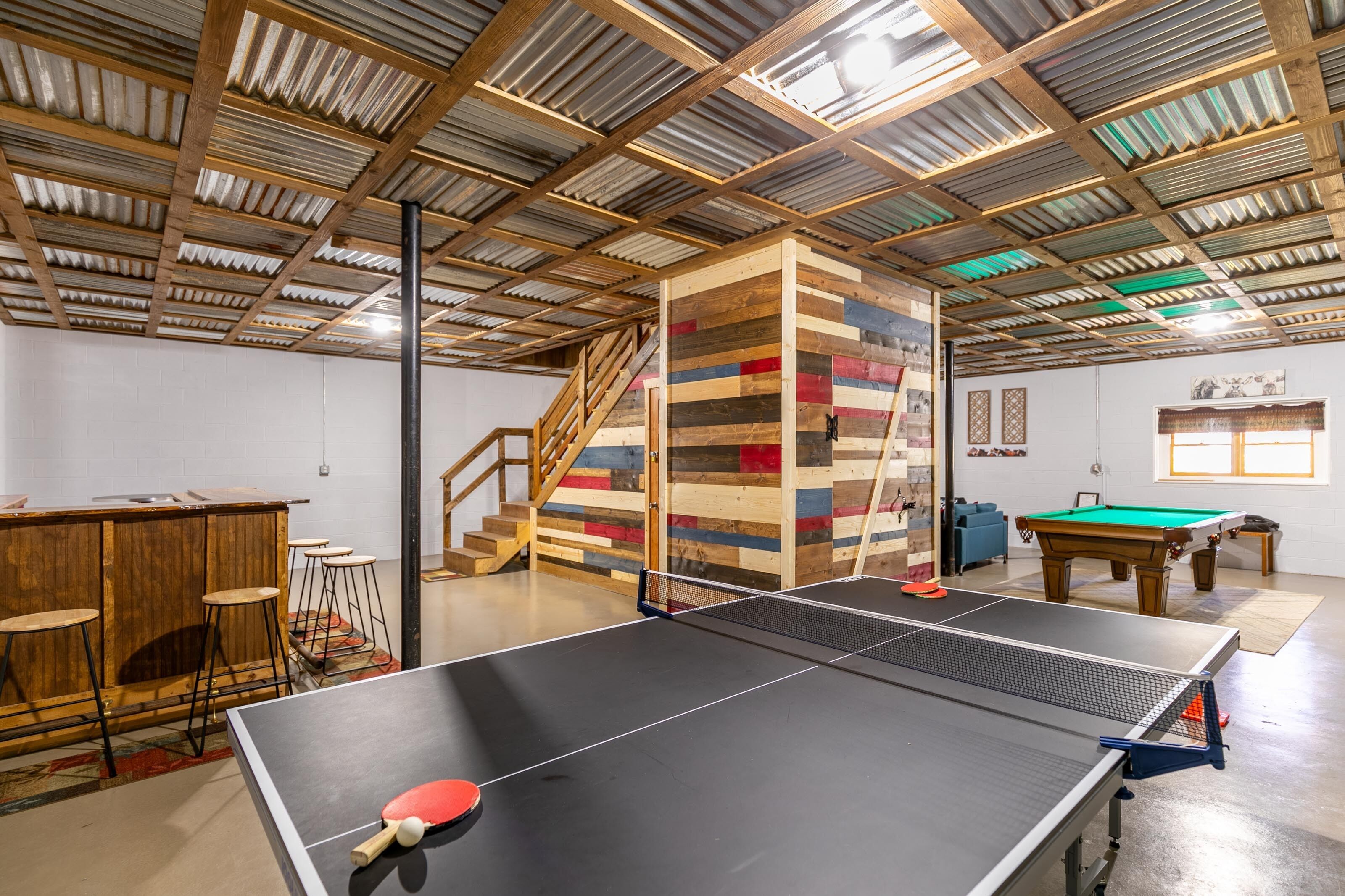 Una mesa de ping pong en una habitación con una barra de madera y taburetes, una mesa de billar y una escalera.