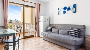 Un salon moderne avec un canapé gris, une table à manger avec des chaises, un réfrigérateur blanc et un balcon offrant une vue sur les palmiers.