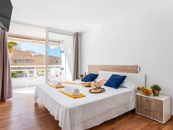 Une chambre d’hôtel moderne avec un grand lit, une tête de lit en bois, une table de chevet sur laquelle se trouve un panier de fruits, et une vue sur la ville grâce à une porte-fenêtre coulissante.