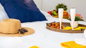 Ein ordentlich bezogenes Bett mit einem blauen Kissen, einem Strohhut, Sonnenbrille und einem Tablett mit Kerzen, Obst und Blumen.