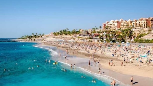 Ein Strand mit badenden und sonnenbadenden Personen, einem klaren blauen Meer und Gebäuden im Hintergrund.