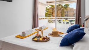 Ein Hotelzimmer mit Bett, einem kleinen Tisch mit Handtüchern und Toilettenartikeln, einem Fenster mit Blick auf Palmen und Gebäude sowie einem Balkon mit Gartenmöbeln.