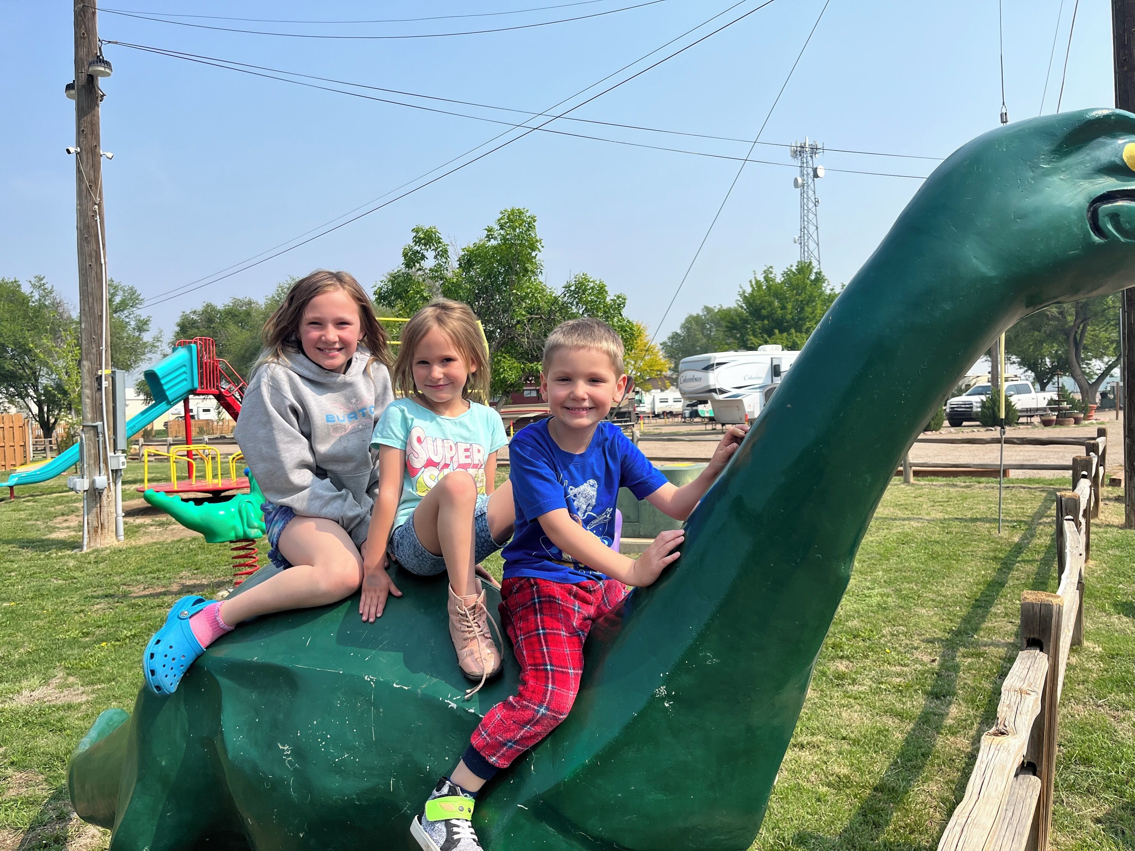 Three children sitting on a green dinosaur-shaped structure in a park.