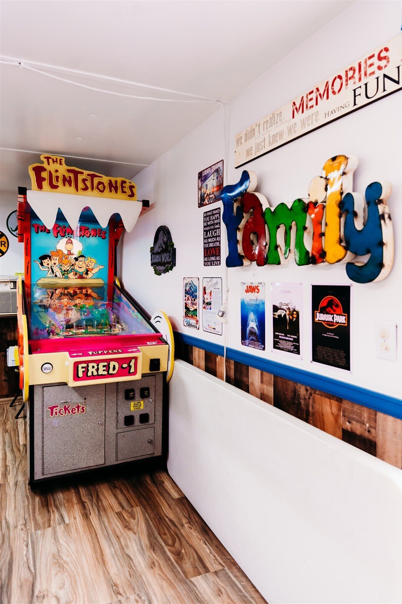 A pinball machine labeled 'The Flintstones' and 'Fred-1' in a room with wooden flooring and a wall decorated with movie posters and a 'Memories Fun' sign.