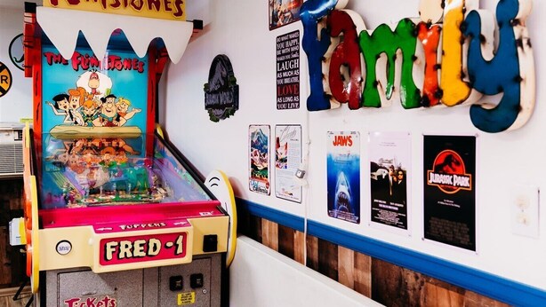 Huoneessa, jossa on puulattia ja seinä, joka on koristeltu elokuvajulisteilla ja jossa on kyltti, jossa lukee ”Memories Fun”, seisoo flipperiautomaatti, jonka kyljessä on teksti ’The Flintstones’ ja ’Fred-1’.