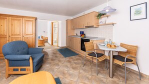 Een compacte keuken met houten kastjes, een eethoek met tafel en stoelen, en een blauwe fauteuil.
