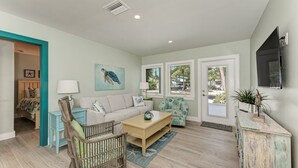 A spacious living room with a light-colored sofa, a wooden coffee table, and a teal accent chair. There is a large window with a view of palm trees and a door leading outside.
