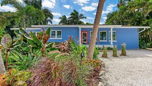A blue house with a red door, surrounded by tropical plants and a gravel driveway.