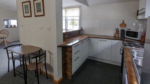 Una cocina moderna con armarios blancos, encimera de madera y estufa integrada. Hay una zona de comedor con una mesa y sillas, y dos cuadros en la pared.
