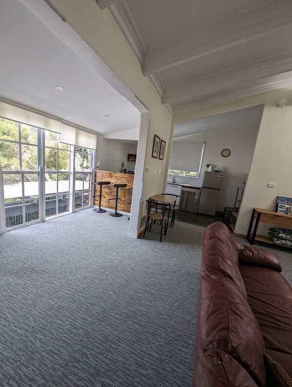 A spacious living area with a large window, a kitchen in the background, and a brown leather sofa.