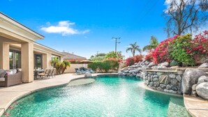 Une piscine au design incurvé, entourée d’un patio aménagé avec du mobilier d’extérieur, un mur en pierre et une végétation luxuriante.
