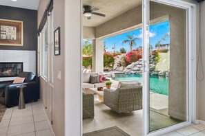 Ein modernes Wohnzimmer mit Kamin, Essbereich und einer Schiebeglastür, die auf eine Außenpatio mit Pool und tropischer Bepflanzung führt.
