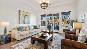 Un salon spacieux avec un canapé beige, une table basse et un fauteuil marron. Une grande fenêtre offre une vue sur des palmiers et un paysage rocheux.