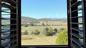 A room with a large window featuring horizontal blinds, a view of a rural landscape with fields and trees, and a simple, functional interior design.