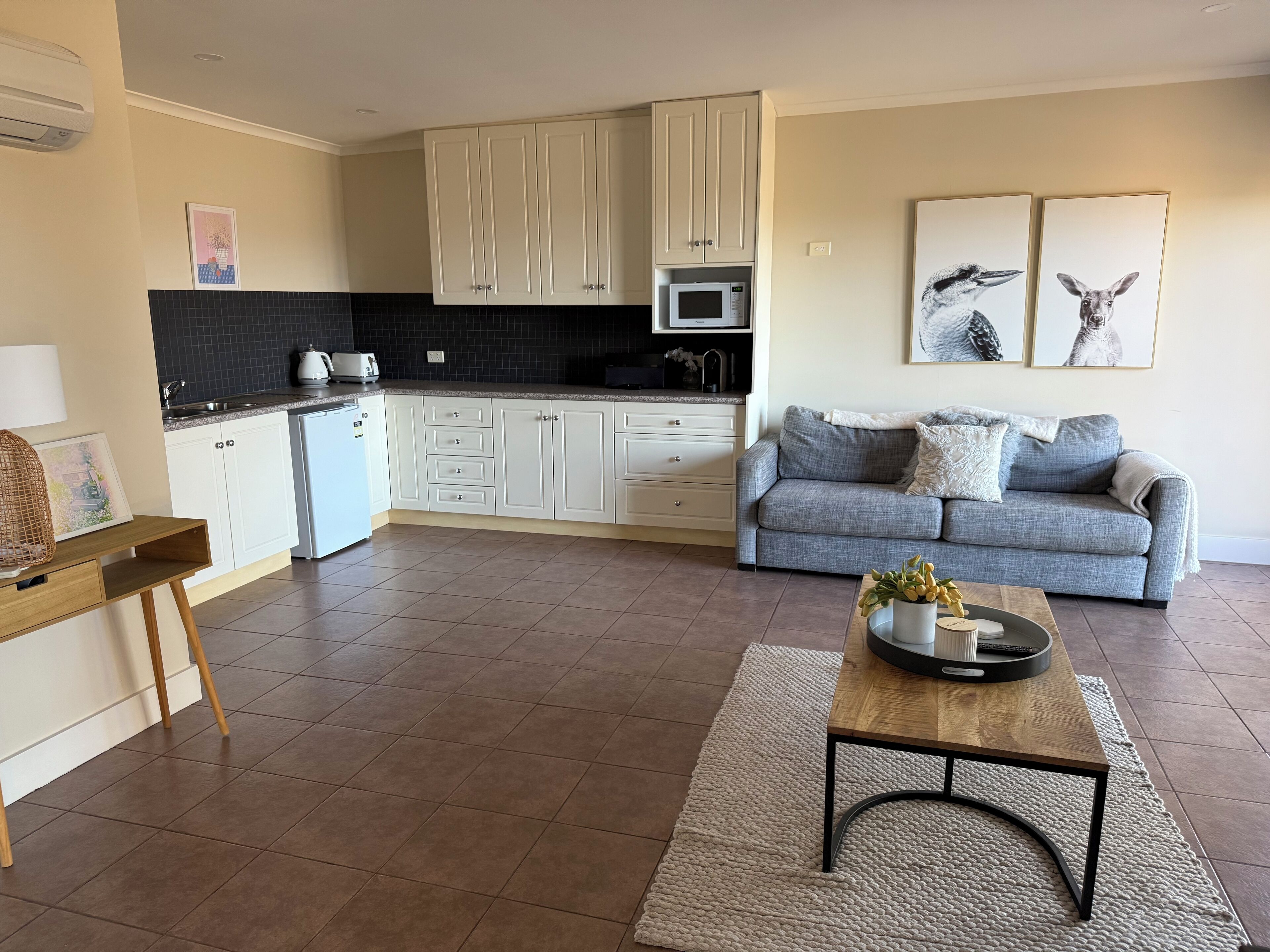Una cocina moderna con armarios blancos, un microondas y un fregadero. En la sala de estar, hay un sofá gris con una alfombra blanca y una mesita de centro.