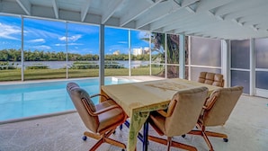 A modern meeting room with a large table, comfortable chairs, and large windows offering a view of a lake and buildings. - Bayfront Duplex! Gulf Access w/ Dock, Pool, Bikes, Kayak & Over $100 of Free Daily Activities! (Bonita Springs)