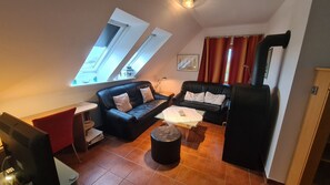 Una sala de estar acogedora con un sofá de cuero negro, una mesita de centro pequeña y un reposapiés negro. Hay dos ventanas con cortinas, un cuadro en la pared y un mueble negro.