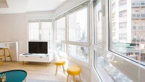A modern living room with a large window, a flat-screen TV, a small table, and two yellow stools. - E 2 BEDROOM FAMILY APARTMENT SAN MARCIAL 33 (San Sebastián)
