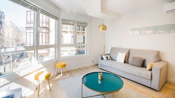 A modern living room with a grey sofa, yellow stools, a teal coffee table, and large windows offering a city view. - E 2 BEDROOM FAMILY APARTMENT SAN MARCIAL 33 (San Sebastián)