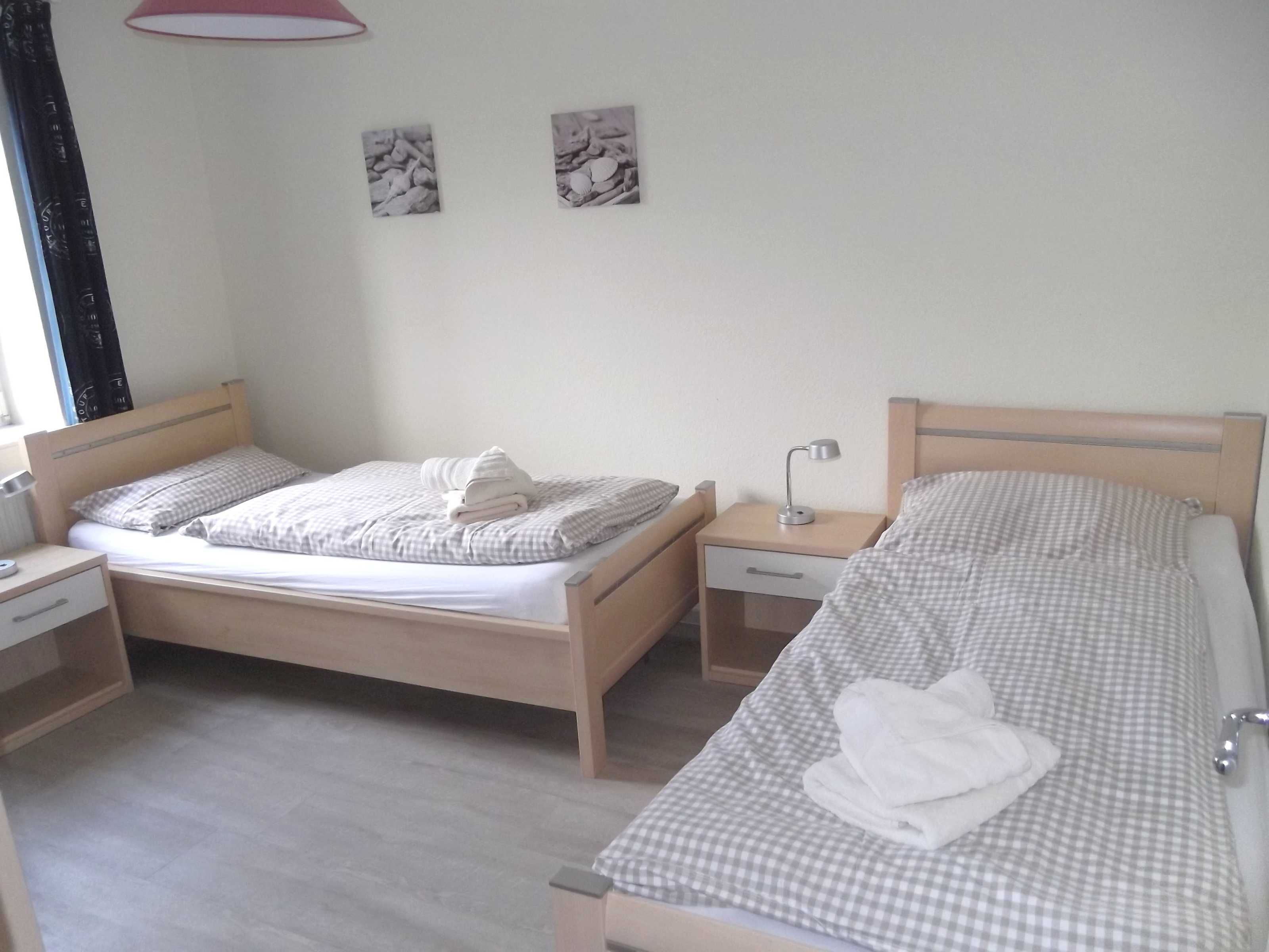 Habitación de hotel con dos camas individuales, cada una con una colcha a cuadros y una toalla doblada. Hay mesitas de noche con lámparas y un cajón pequeño. El piso es de color claro y la pared es lisa con dos cuadros enmarcados.