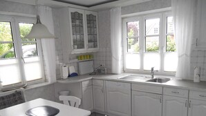 A kitchen with white cabinets, a sink, a window with curtains, and a table with chairs. - Haferweg 2, Strandnelke EG - Haferweg 2, Strandnelke (Wyk auf Föhr)